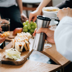Person holding stainless steel travel coffee grinder at table with bread, grapes, and beverages during social gathering