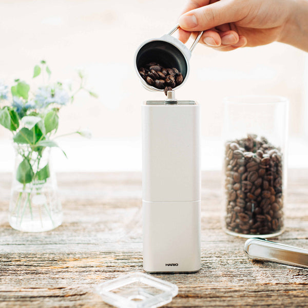 Hand pouring roasted coffee beans from metal measuring cup into white Hario grinder