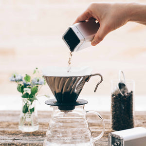 Pouring ground coffee from white ceramic burr grinder into glass carafe with filter