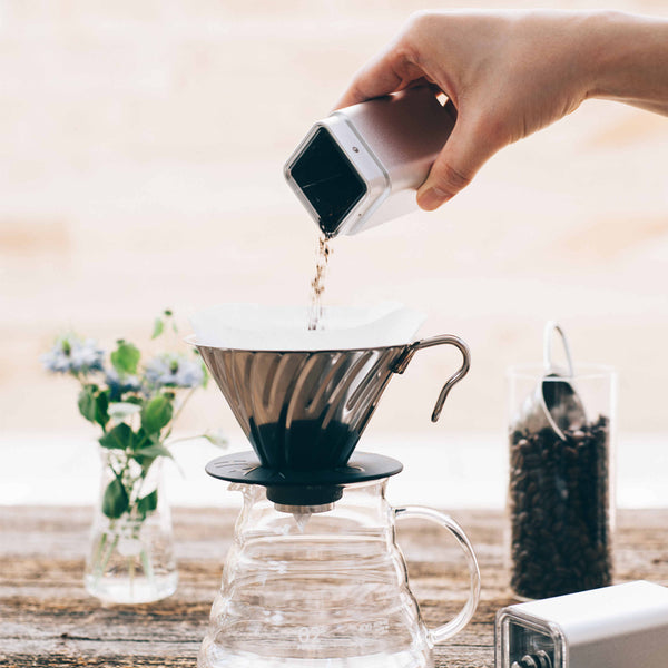 Pouring ground coffee from white ceramic burr grinder into glass carafe with filter