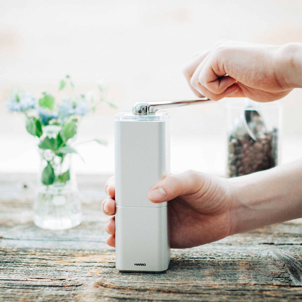 Person using white Hario Prism ceramic burr grinder with metal spoon over jar