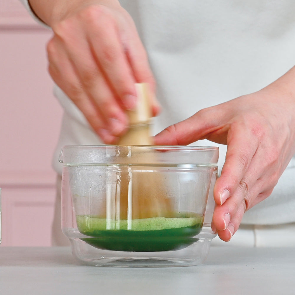 Hands stirring matcha with wooden whisk in clear glass bowl on white surface.