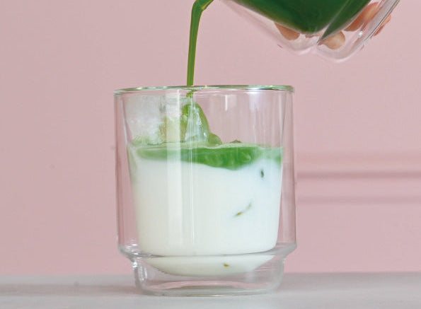 Green matcha tea being poured from glass pitcher into double-walled cup with milk base and fresh mint leaf.