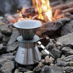 Hario V60 metal coffee dripper and server positioned on rocks beside a campfire with flames