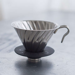 Stainless steel V60 dripper with ribbed cone interior and metal handle on marble surface.
