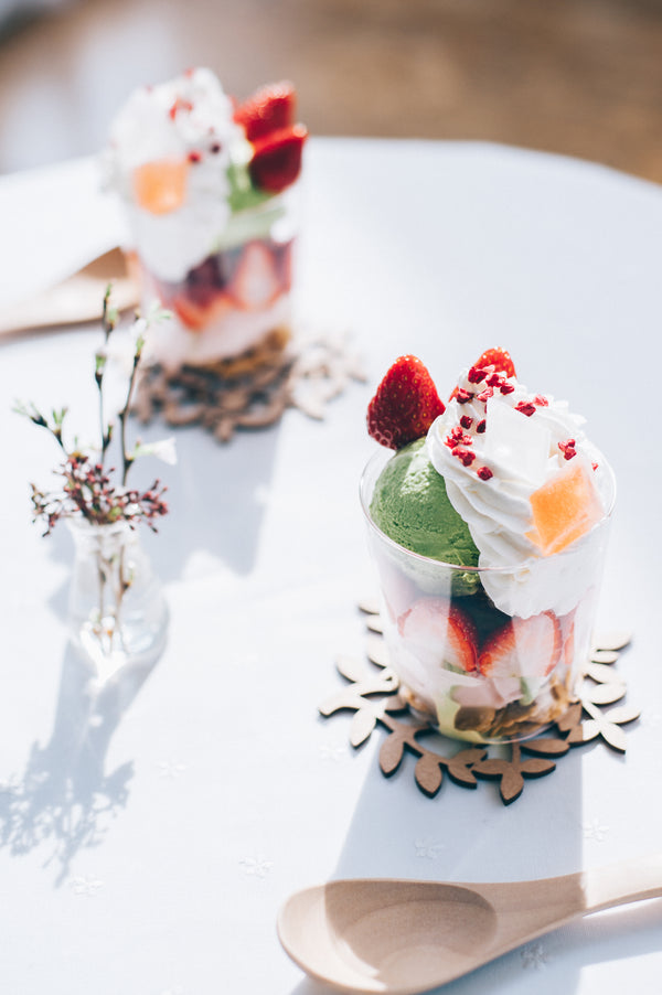 Decorated glass dessert with pistachio ice cream and strawberry beside fresh flowers in vase.