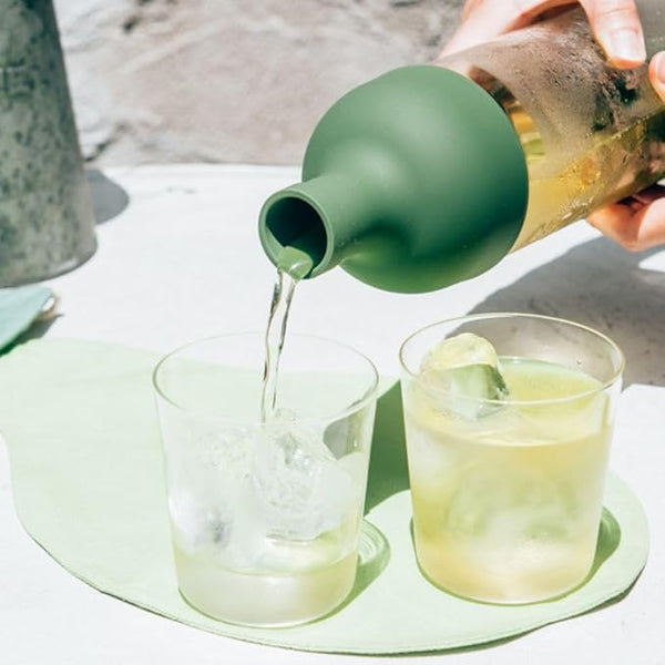 Green ceramic pitcher pours pale yellow beverage into two clear glasses with ice.