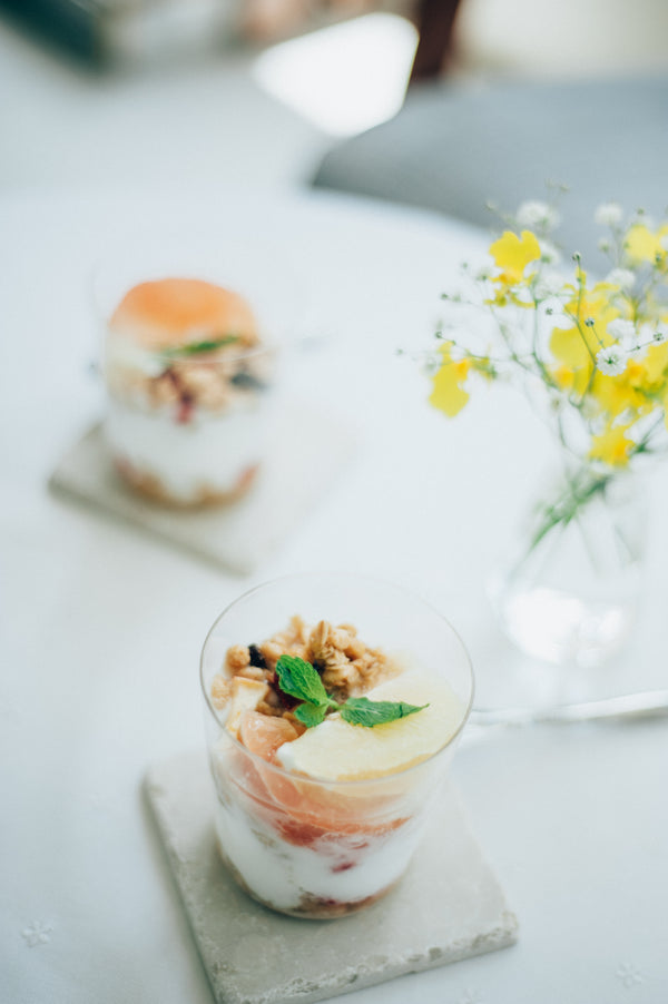 Clear glass dessert with pink base layer, cream, granola, grapefruit slice, and mint on concrete coaster.
