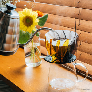 Gooseneck kettle pouring water into yellow Suiren dripper over clear glass carafe.