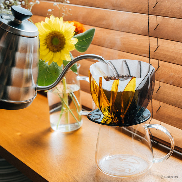 Gooseneck kettle pouring water into yellow Suiren dripper over clear glass carafe.