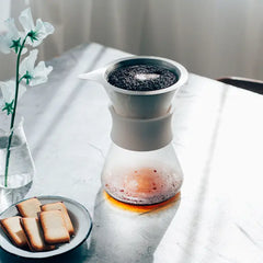 Hario glass coffee maker with white mesh filter top, filled dark coffee below, beside toast and white flowers.
