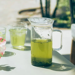 Glass pitcher with pale yellow-green herbal tea and floating herb sprigs in outdoor sunlight.