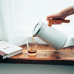Person pouring dark coffee from white stainless steel Hario thermal pot into glass cup on wooden desk.