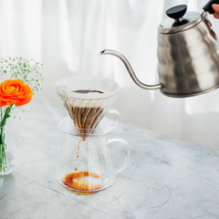 Stainless steel gooseneck kettle pouring hot water into glass V60 dripper with coffee filter over carafe.