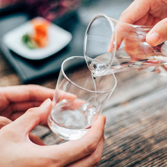 Person pouring water into clear glass over another glass on wooden table with plated food blurred in background