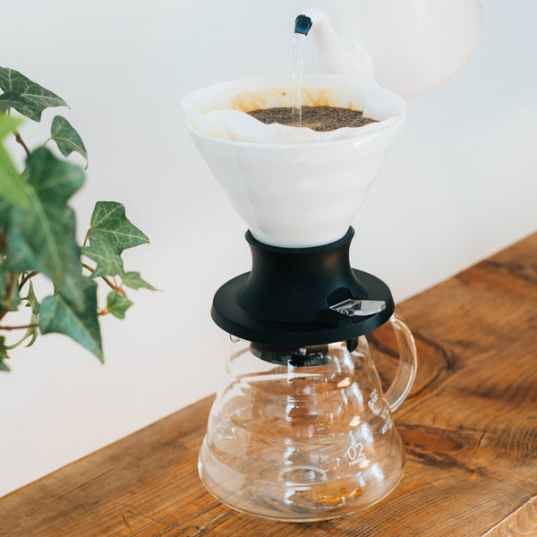 White ceramic V60 dripper with black base actively brewing coffee over glass carafe on wooden table.