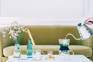 Hands pouring hot water into Largo dripper tea set on white coffee table in bright interior.