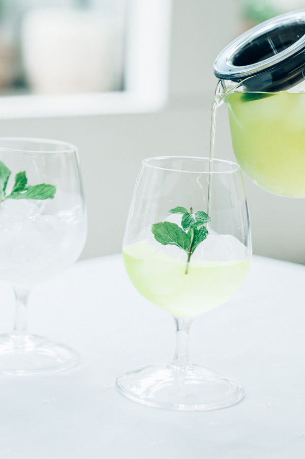 Pale yellow tea being poured from black-topped Largo dripper into glass carafe with mint garnish.