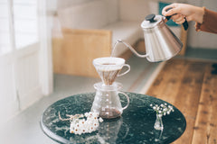 Person pouring hot water from gooseneck kettle into V60 dripper cone over coffee grounds on dark green table with white