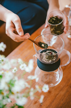 Hands adding loose tea leaves with wooden spoon into glass cup during tea preparation ritual.