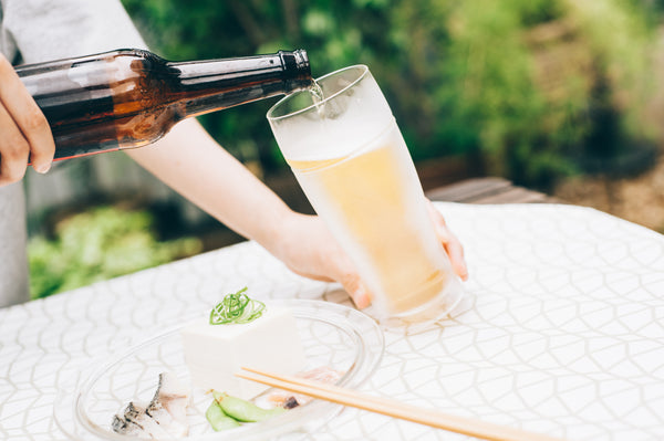 Dark beer bottle pouring golden liquid into tall glass on outdoor white patterned table