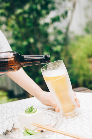 Brown bottle dispensing pale ale into double-walled glass against blurred green garden