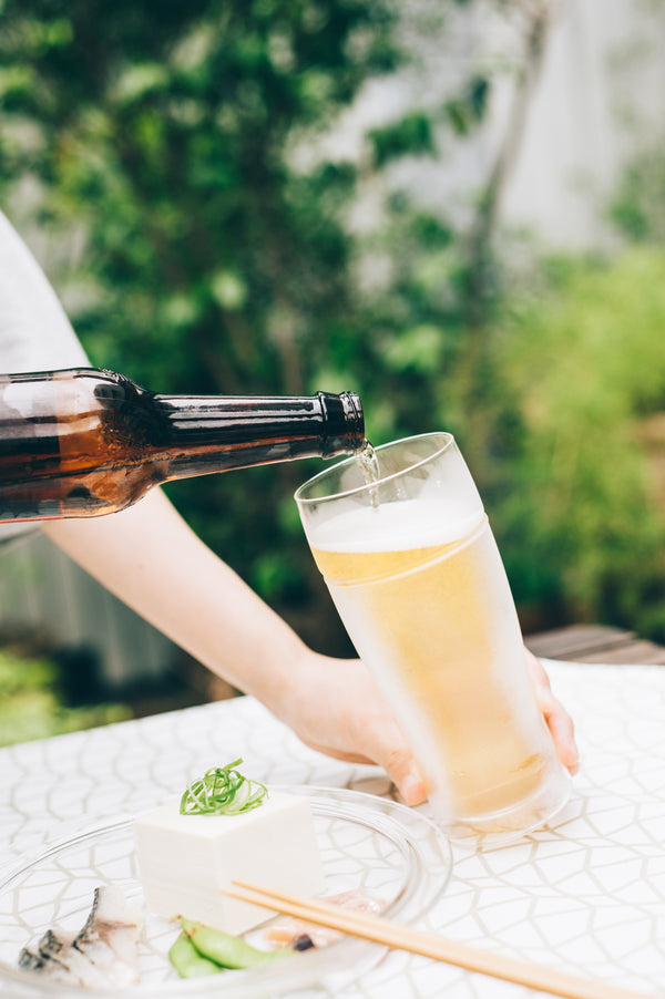 Brown bottle dispensing pale ale into double-walled glass against blurred green garden