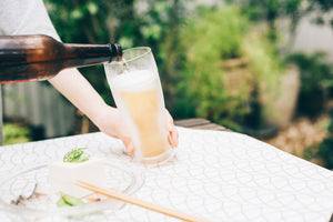 Beer bottle tilted mid-pour into tall glass on white tablecloth with food plate