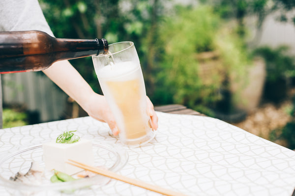 Beer bottle tilted mid-pour into tall glass on white tablecloth with food plate