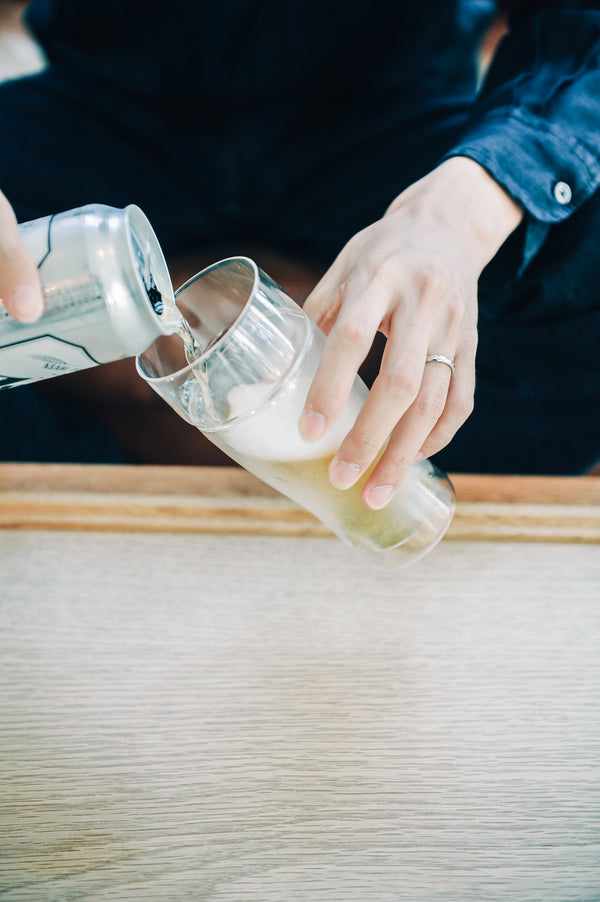 Person in denim pouring pale liquid from measuring cup into double-walled glass on wooden table