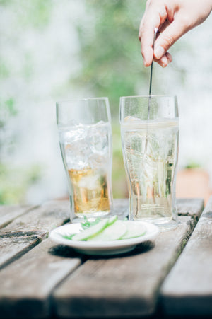 Hand stirring ice in clear sparkling water inside double-walled glass on rustic wooden table with cucumber slices on white