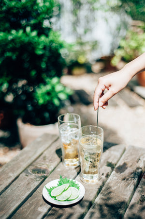 Person inserting green drink stirrer into tall double-walled glass filled with clear beverage and ice in sunny garden setting