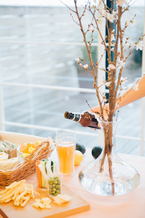 Overhead view of orange juice in tall glass beside bread basket, vegetables, and dried branches in vase with ocean view
