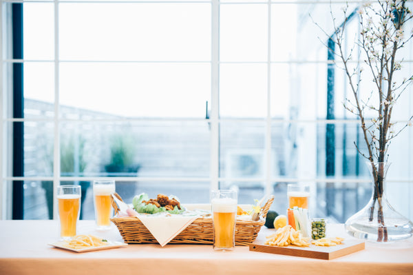 Four glasses of golden beer with white foam held by multiple hands in celebratory toast over food-filled table indoors
