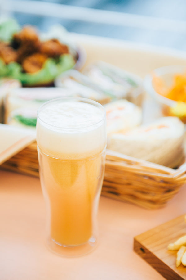 Golden beer with white foam head in tall glass beside wooden serving board with appetizers and beach backdrop