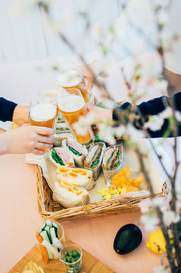 Multiple hands clinking tall glasses filled with pale ale and sandwiches arranged in wicker basket on table