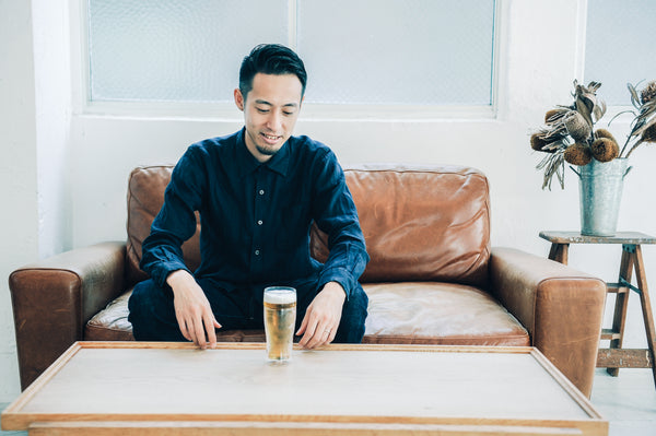 Man in blue shirt smiling while holding beer glass on brown leather sofa near window