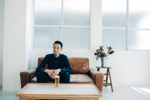 Seated man in navy shirt with beer glass on wooden table, large windows behind