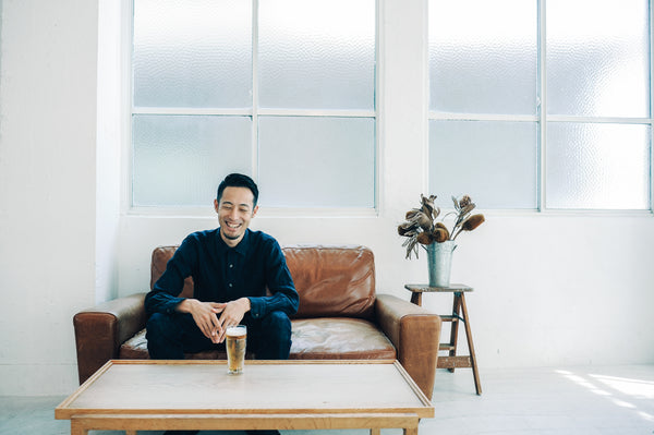Man in dark blue button-up sitting on brown couch holding tall beer glass indoors