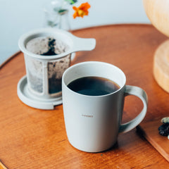 White ceramic mug filled with black coffee on wooden table with mesh filter infuser in background