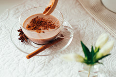 Clear glass teacup filled with chocolate beverage topped with cocoa powder, accompanied by star anise and cinnamon stick