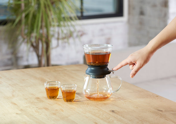 Hands removing full amber tea dripper cup from glass server base on wooden table.