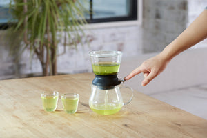 Hands lifting pale green tea-filled Largo dripper above matching glass server on wooden surface.
