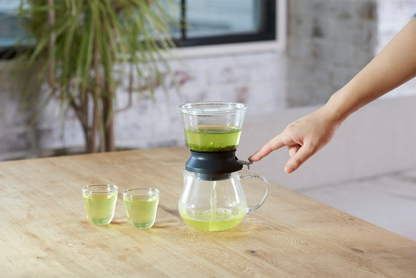 Hands lifting pale green tea-filled Largo dripper above matching glass server on wooden surface.