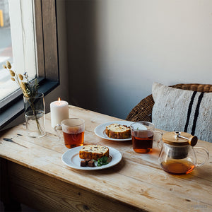 Wooden table setting with two plates of sliced cake, lit candle, and glass teapot with wooden lid