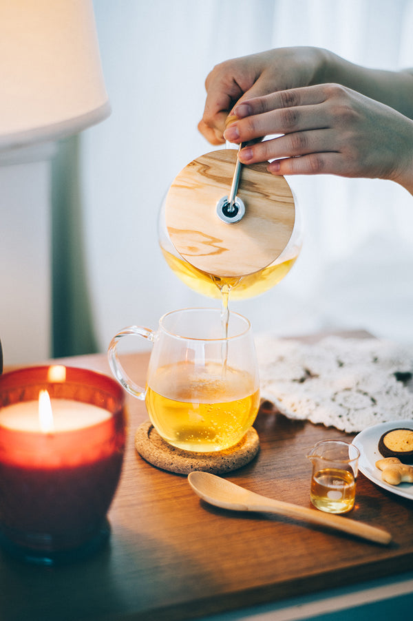 Hands pouring golden tea from olive wood lid into clear glass mug