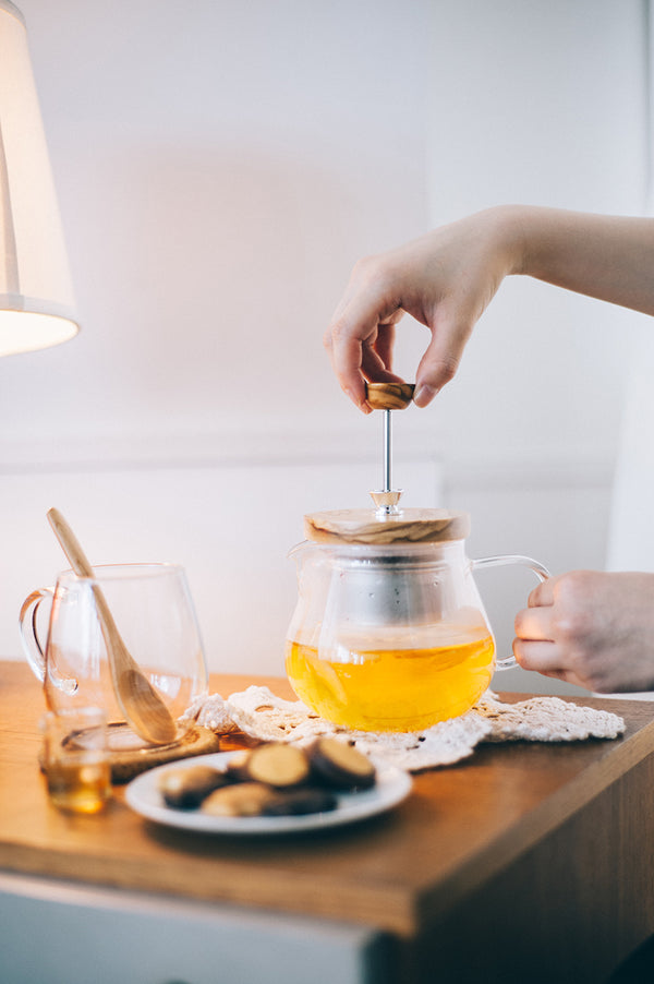 Hand lifting wooden tea infuser lid above glass teapot filled with yellow-golden tea