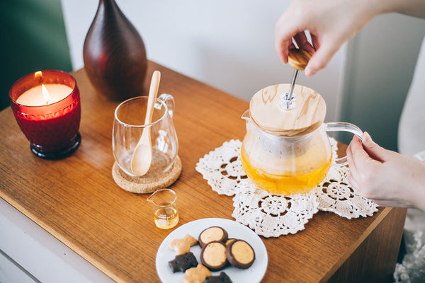 Overhead view of wooden table with glass teapot, cookies on plate, red candle, and tea accessories