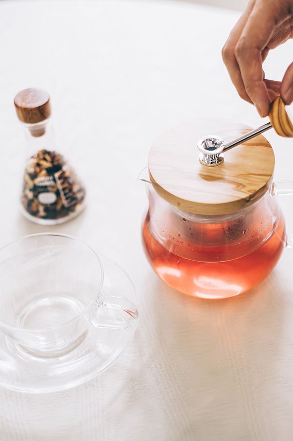 Hands placing wooden lid on glass teapot containing brewed reddish tea
