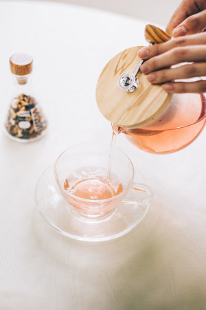 Pouring amber-colored tea from wooden-lidded pot into clear handleless cup